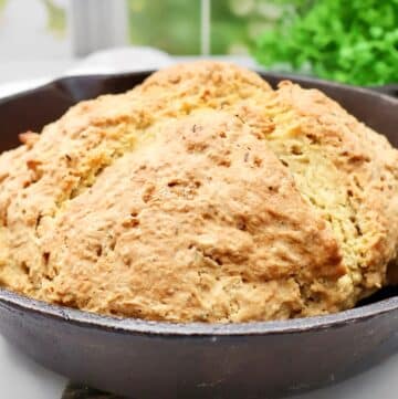 A round, golden-brown loaf of Irish soda bread with caraway sits in a black cast iron skillet, its textured, rustic crust inviting. Green leafy plants and a window are softly blurred in the background.