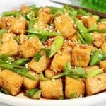 A plate of crispy golden tofu cubes topped with chopped green onions, sesame seeds, and a drizzle of sauce, served on a white dish.