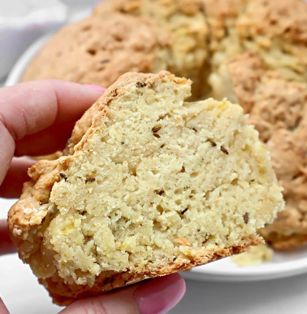A hand holds a thick slice of crumbly, golden-brown Irish soda bread with caraway, showing its dense, textured interior with visible seeds and bits of cheese or potato. The rest of the loaf is in the background.