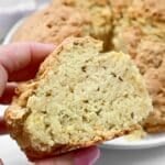 A hand holds a thick slice of crumbly, golden-brown Irish soda bread with caraway, showing its dense, textured interior with visible seeds and bits of cheese or potato. The rest of the loaf is in the background.
