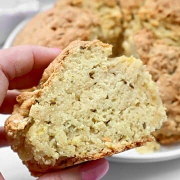 A hand holds a thick slice of crumbly, golden-brown Irish soda bread with caraway, showing its dense, textured interior with visible seeds and bits of cheese or potato. The rest of the loaf is in the background.