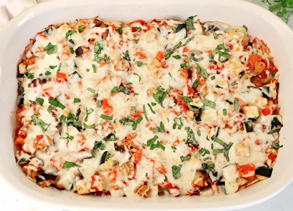 A casserole dish filled with baked ratatouille topped with melted cheese and chopped fresh herbs, showing colorful pieces of vegetables like tomatoes, zucchini, and eggplant.
