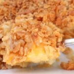 A close-up of a cheesy Southern side dish casserole, topped with a golden, crispy cracker and breadcrumb crust, with melted cheese and potato chunks visible underneath—perfect for those who love classic pineapple casserole flavors.