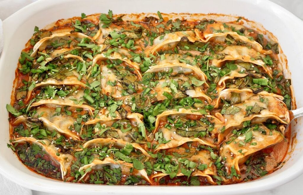 A white baking dish filled with dumplings in a red sauce, topped with chopped fresh herbs and sesame seeds. The dumplings are arranged in neat rows and slightly browned on the edges.