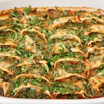 A white baking dish filled with dumplings in a red sauce, topped with chopped fresh herbs and sesame seeds. The dumplings are arranged in neat rows and slightly browned on the edges.