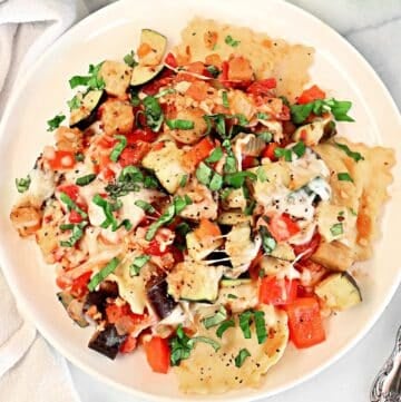 A plate of ravioli topped with roasted vegetables like eggplant and red bell pepper, garnished with chopped fresh basil and melted cheese.