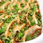 A close-up of a white dish filled with neatly arranged pan-fried dumplings, garnished with chopped fresh cilantro, green onions, and sprinkled with sesame seeds.