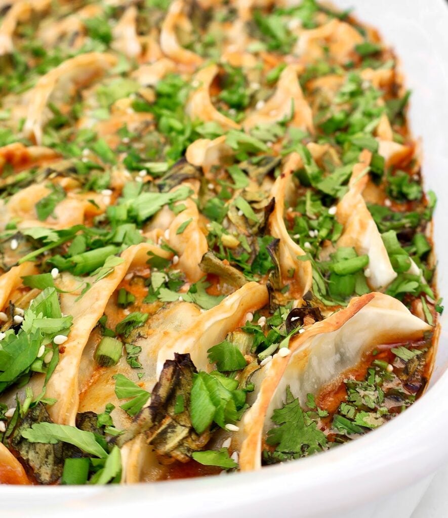 A close-up of a white dish filled with neatly arranged pan-fried dumplings, garnished with chopped fresh cilantro, green onions, and sprinkled with sesame seeds.