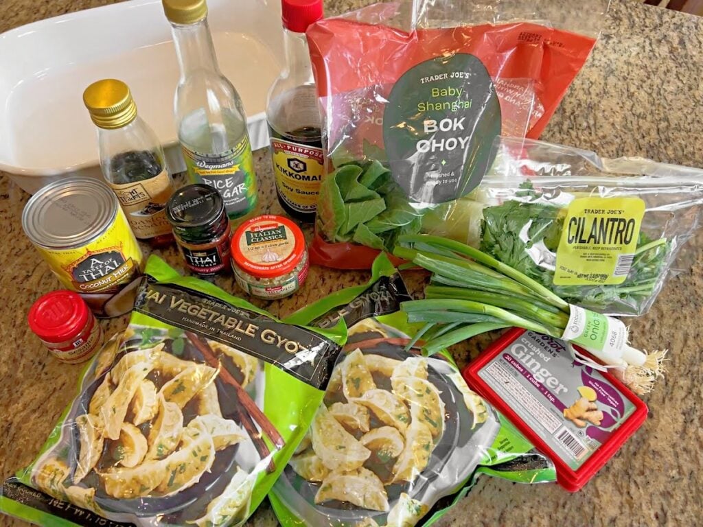 Assorted Asian-inspired ingredients on a kitchen counter, including bok choy, cilantro, green onions, ginger paste, soy sauce, rice vinegar, canned water chestnuts, chili paste, and bags of vegetable gyoza.