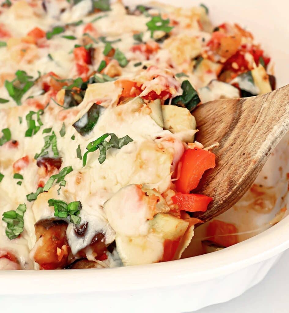 A wooden spoon scoops a serving of cheesy baked vegetable casserole from a white dish, showing colorful pieces of zucchini, red bell pepper, tomato, and fresh herbs on top.