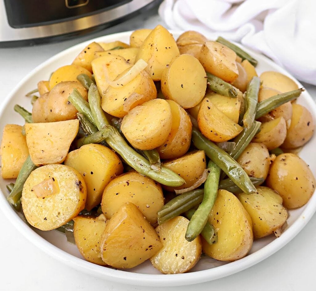 A white bowl filled with cooked, seasoned baby potatoes and green beans, with visible bits of onion—just like a homemade Southern green beans recipe. The vegetables are cut into chunks and lightly peppered, set against a backdrop of a kitchen appliance and white cloth.