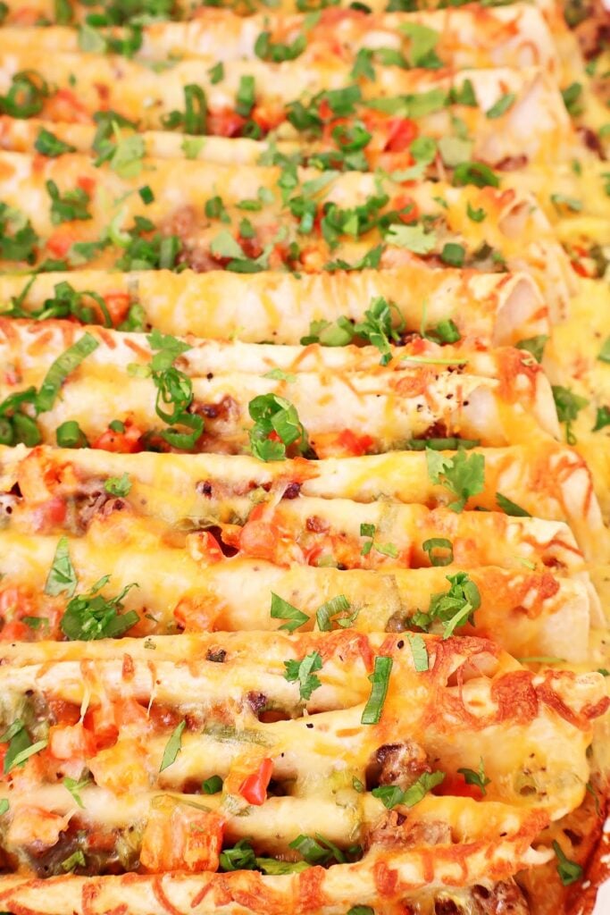 Cheesy baked enchiladas topped with chopped tomatoes, green onions, and cilantro, arranged closely together in a pan—this dish is the perfect easy Mexican breakfast casserole for your next brunch or family gathering.