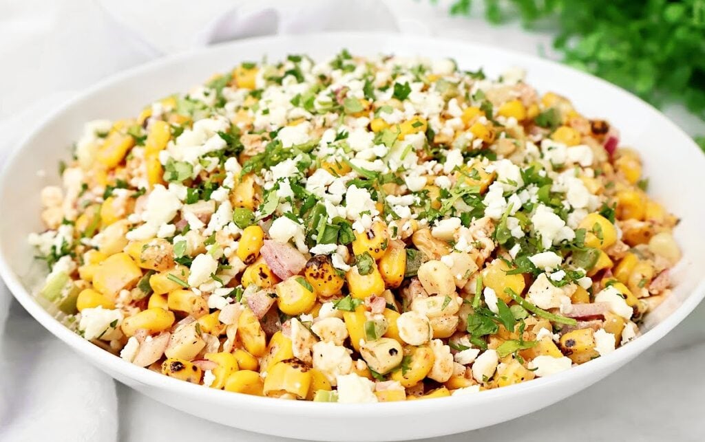 A white plate filled with a colorful corn salad, featuring grilled corn, diced red onion, peppers, chopped cilantro, and crumbled cheese, garnished with fresh herbs.
