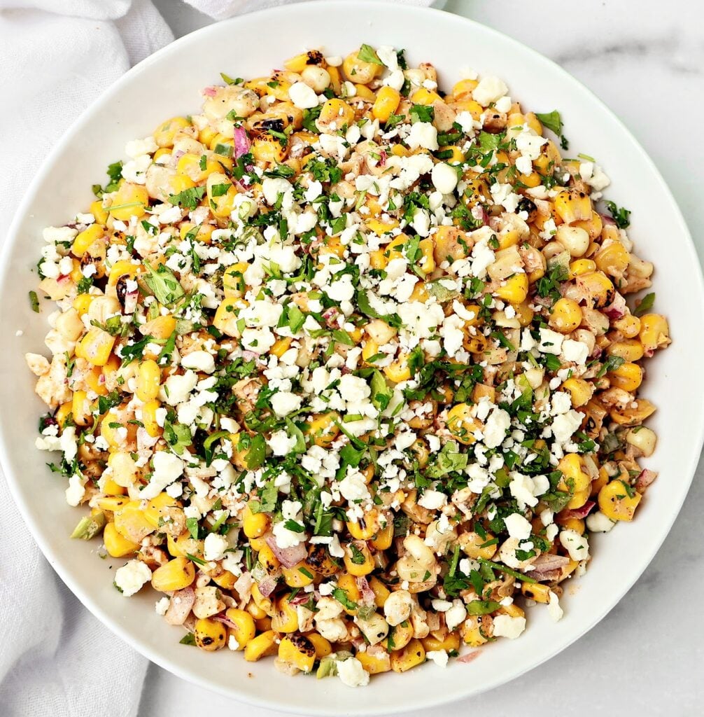 A white bowl filled with Mexican street corn salad topped with crumbled cheese, chopped cilantro, and a mix of corn, red onion, and green peppers on a marble surface.