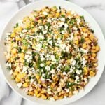 A white bowl filled with Mexican street corn salad topped with crumbled cheese, chopped cilantro, and a mix of corn, red onion, and green peppers on a marble surface.