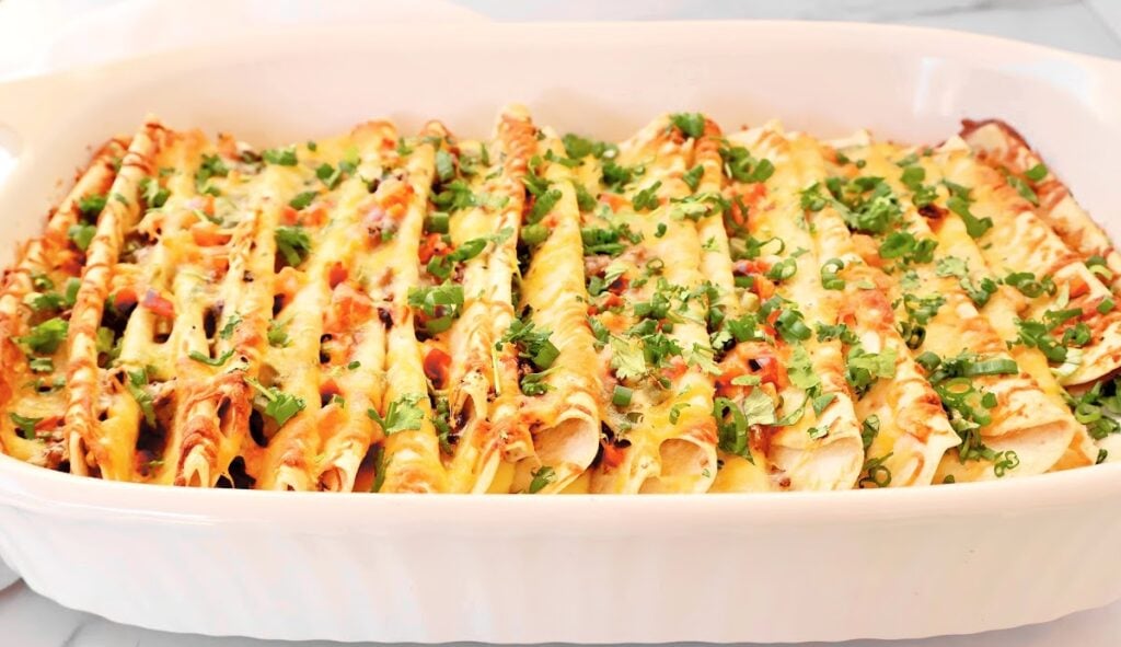 A white baking dish filled with neatly arranged easy Mexican breakfast casserole enchiladas, topped with melted cheese and garnished with chopped green herbs.