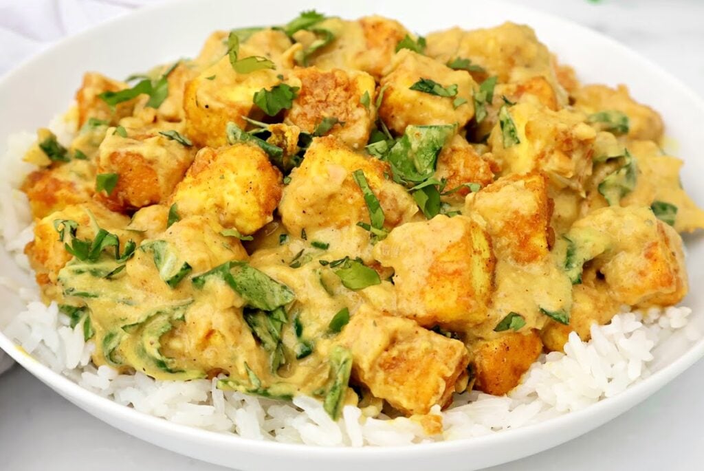 A white bowl filled with white rice topped with creamy tofu curry, garnished with chopped fresh cilantro and spinach. The tofu cubes are coated in a yellow sauce.