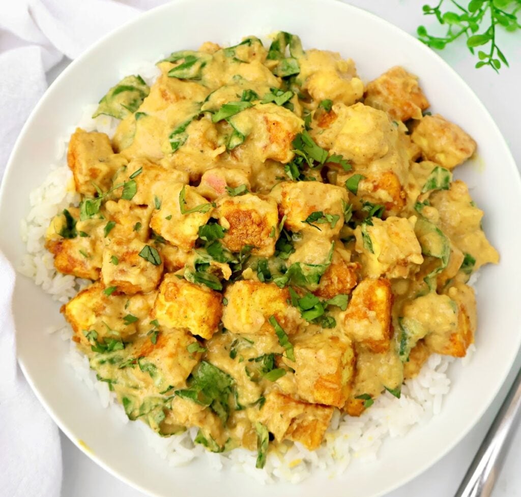 A white plate filled with white rice topped with golden-brown tofu cubes and a creamy spinach curry sauce, garnished with chopped parsley. A fork and a green plant are visible nearby.