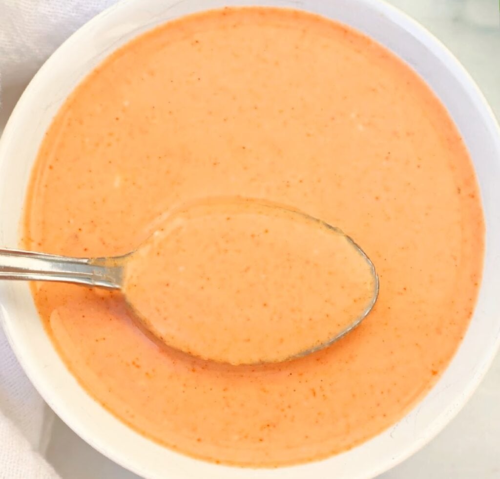 A bowl of creamy Thai peanut sauce with a spoon resting inside, placed on a white surface.