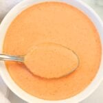 A bowl of creamy Thai peanut sauce with a spoon resting inside, placed on a white surface.