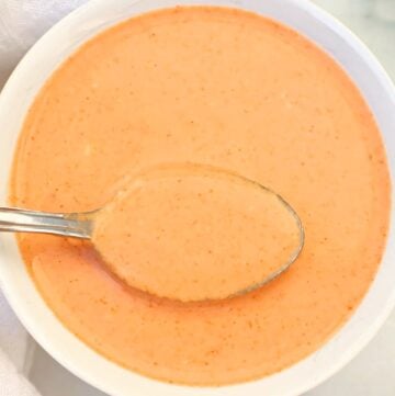 A bowl of creamy Thai peanut sauce with a spoon resting inside, placed on a white surface.