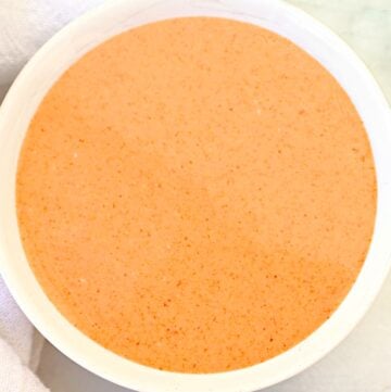 A white bowl filled with a creamy, orange-pink Thai peanut sauce sits on a white surface, with part of a white cloth visible in the corner.