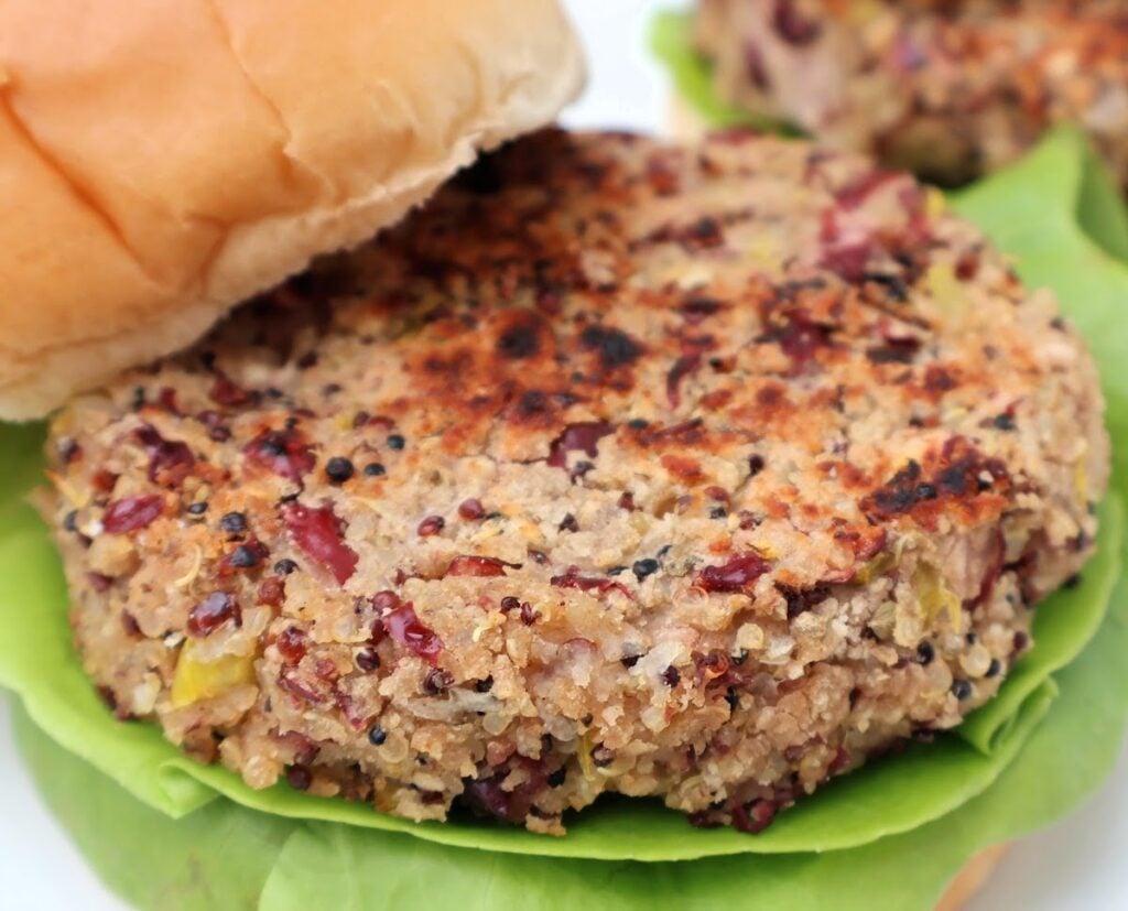 A close-up of a veggie burger patty on a leaf of lettuce, served in a soft bun. The patty has a textured, grainy appearance with visible bits of vegetables and grains.