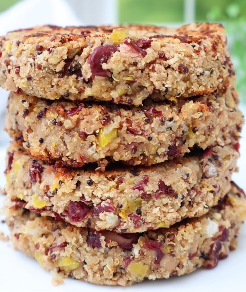 Four thick, round veggie patties stacked on top of each other, showing a textured mix of grains, beans, seeds, and chopped vegetables, with a golden-brown, crispy surface.
