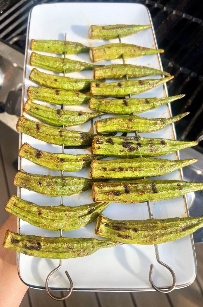 Several Smoky Grilled Okra pods are skewered and arranged neatly on a rectangular white platter, showcasing perfect grill marks. The background features part of a grill, highlighting this classic Southern okra recipe.
