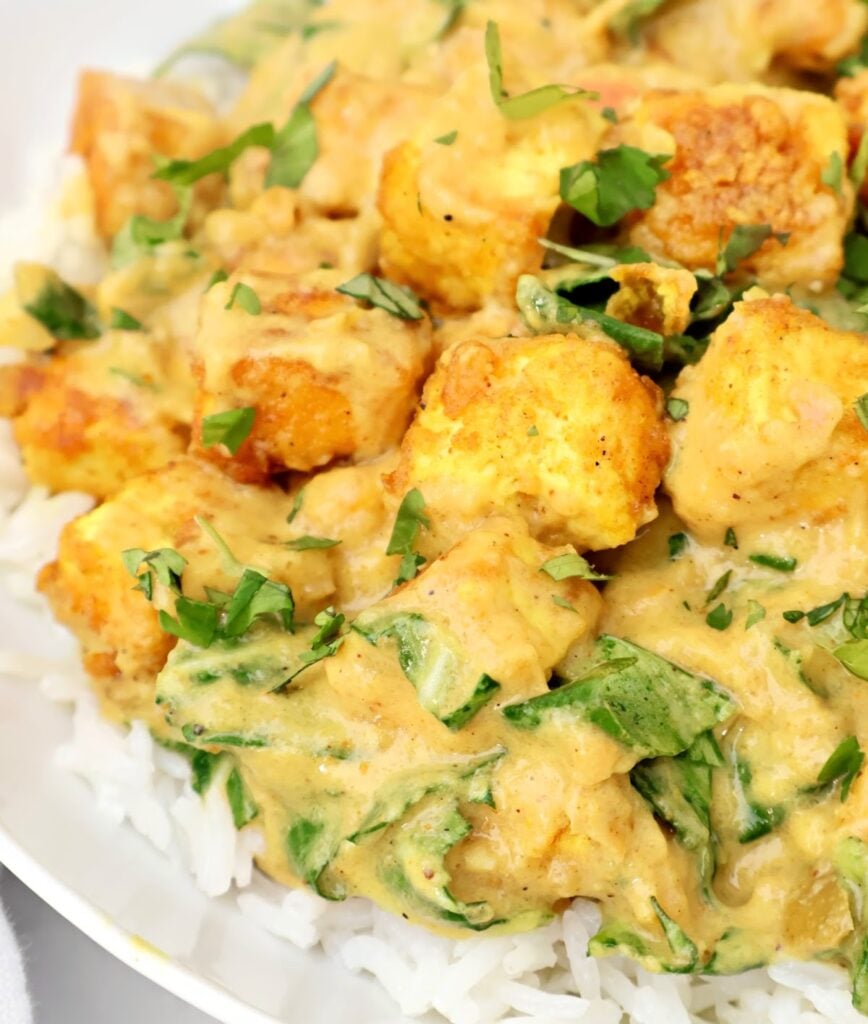 A close-up of golden cubes of tofu in a creamy yellow curry sauce with spinach, served over a bed of white rice and garnished with fresh chopped herbs.