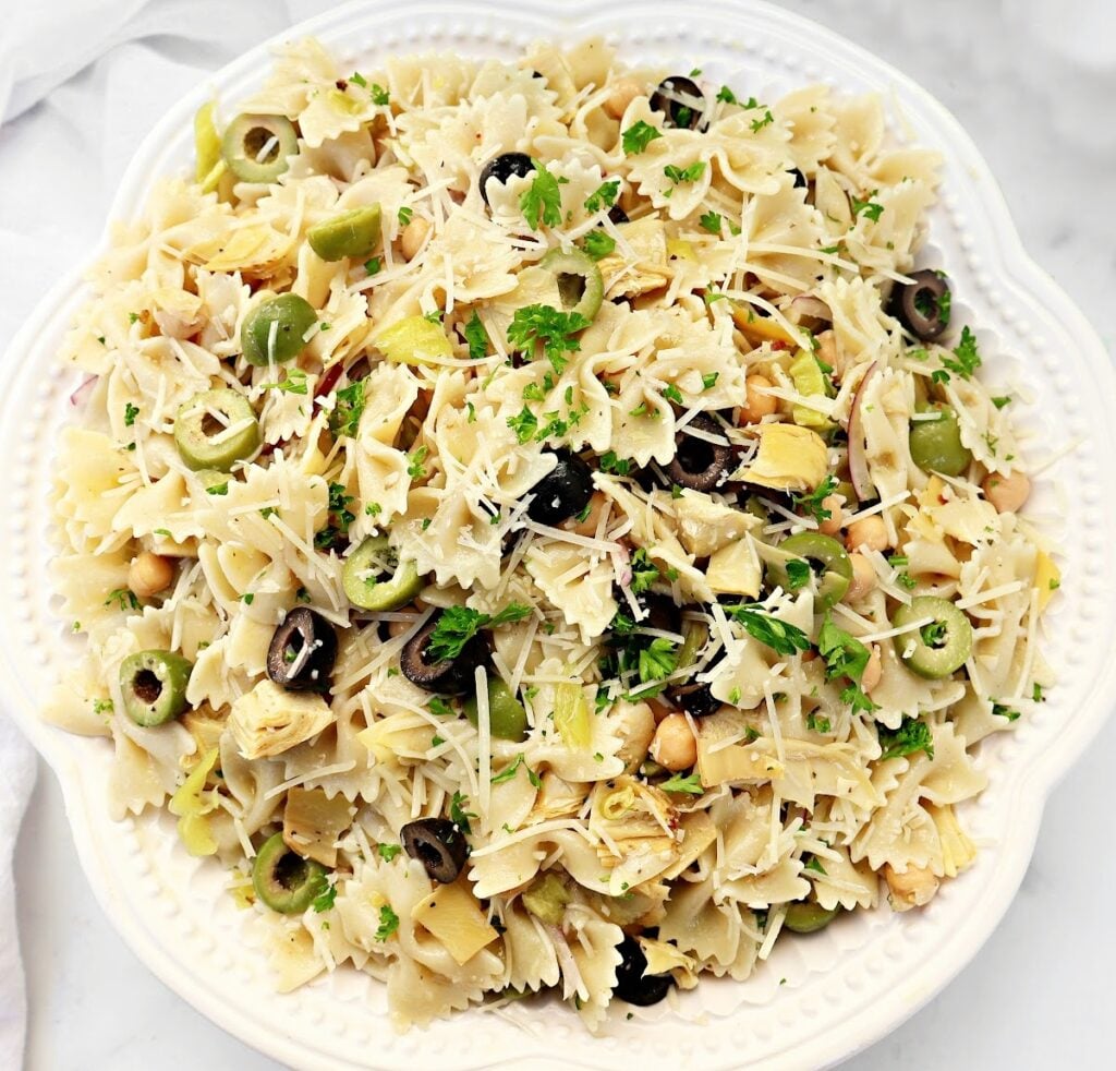 A white plate filled with bowtie pasta salad, garnished with chopped parsley, sliced black and green olives, artichoke hearts, chickpeas, and shredded parmesan cheese.