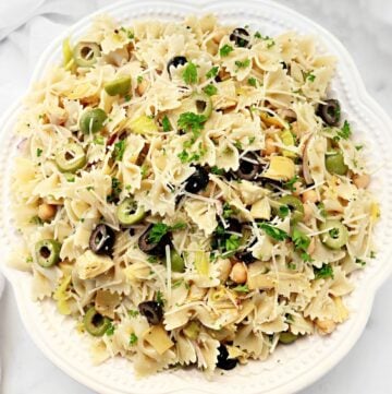 A white plate filled with bowtie pasta salad, garnished with chopped parsley, sliced black and green olives, artichoke hearts, chickpeas, and shredded parmesan cheese.