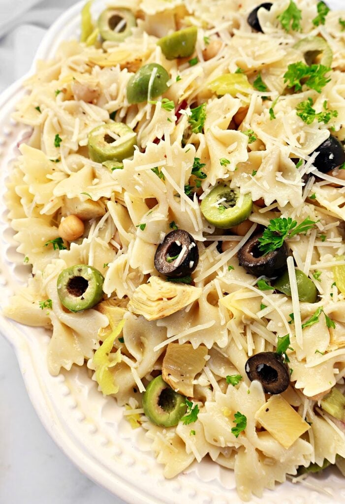 A close-up of bowtie pasta salad on a white plate, topped with sliced green and black olives, artichoke hearts, grated Parmesan cheese, chickpeas, and fresh parsley.