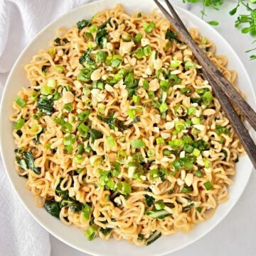 A bowl of cooked bok choy ramen noodles topped with chopped green onions, peanuts, and leafy greens, with a pair of brown chopsticks resting on the side of the bowl.
