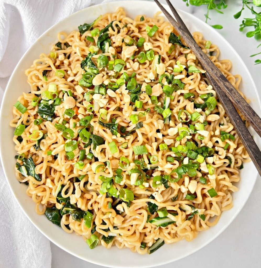 A bowl of cooked bok choy ramen noodles topped with chopped green onions, peanuts, and leafy greens, with a pair of brown chopsticks resting on the side of the bowl.