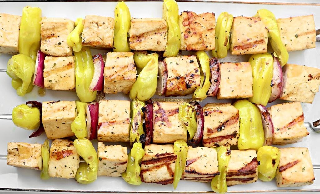 Three grilled skewers with cubed Mississippi Tofu, yellow pepper slices, and red onion pieces, arranged neatly on a white platter. The tofu is lightly charred, and the vegetables are bright and fresh-looking.