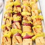 Three metal skewers hold grilled Mississippi Tofu cubes, red onion pieces, and yellow pepper slices, arranged neatly on a white rectangular plate. The tofu has visible grill marks.