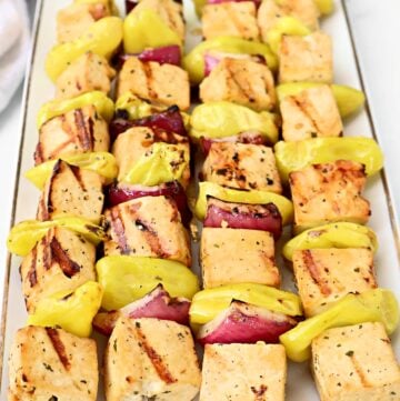 Three metal skewers hold grilled Mississippi Tofu cubes, red onion pieces, and yellow pepper slices, arranged neatly on a white rectangular plate. The tofu has visible grill marks.