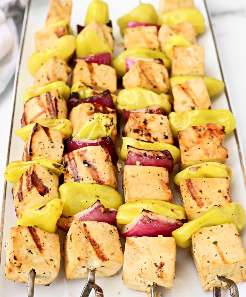 Three metal skewers hold grilled Mississippi Tofu cubes, red onion pieces, and yellow pepper slices, arranged neatly on a white rectangular plate. The tofu has visible grill marks.