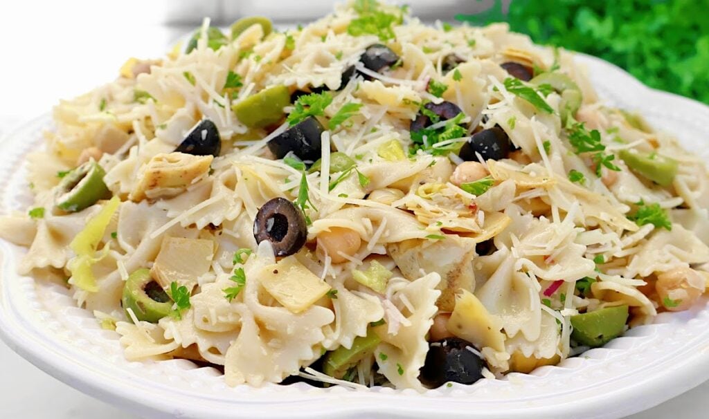 A plate of bowtie pasta salad topped with shredded cheese, sliced black olives, chopped green olives, parsley, and artichoke hearts, served on a white dish with a garnish in the background.