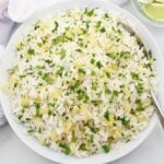 A white bowl filled with fluffy white rice mixed with finely chopped herbs and small pieces of yellow ginger, with a spoon on the side. Lime wedges are visible in a small dish nearby.