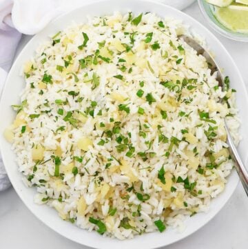 A white bowl filled with fluffy white rice mixed with finely chopped herbs and small pieces of yellow ginger, with a spoon on the side. Lime wedges are visible in a small dish nearby.