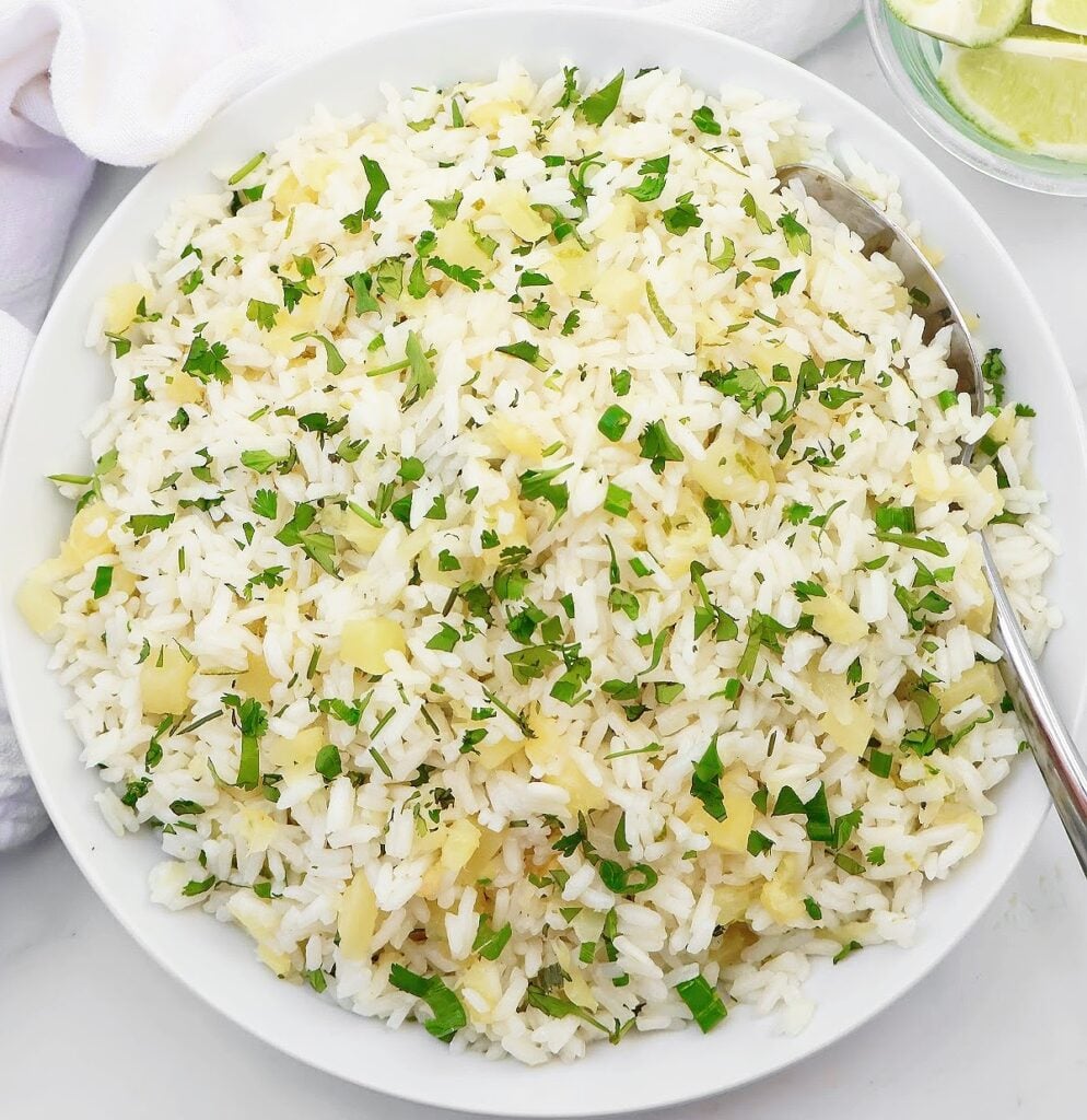 A white bowl filled with fluffy white rice mixed with finely chopped herbs and small pieces of yellow ginger, with a spoon on the side. Lime wedges are visible in a small dish nearby.
