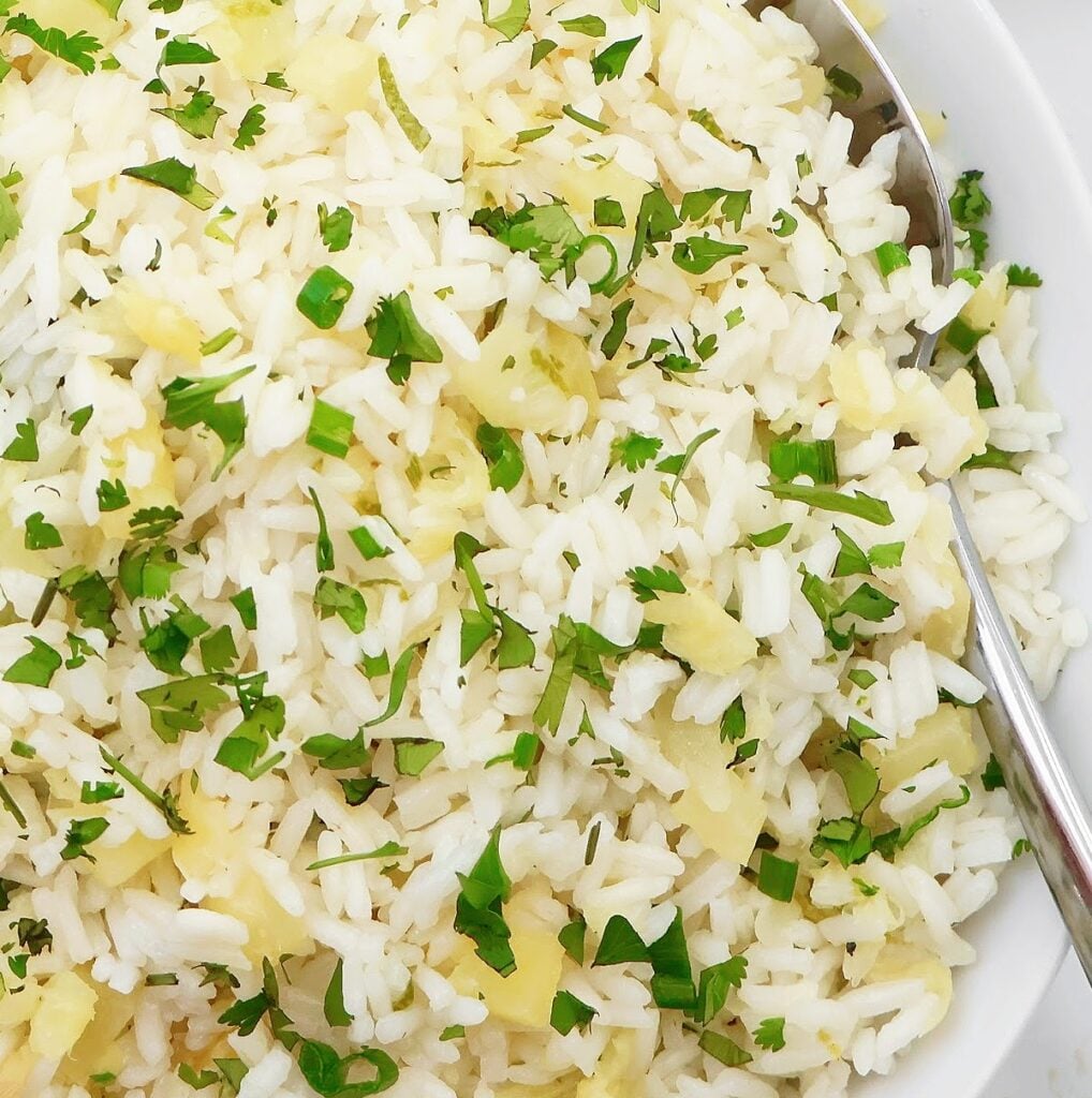 A close-up of a bowl of cooked white rice mixed with finely chopped cilantro and sautéed onions, with a metal spoon on the side.