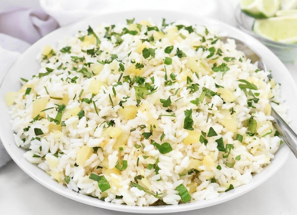 A white bowl filled with white rice mixed with small pineapple pieces and sprinkled with chopped fresh herbs, with a spoon on the side. Lime wedges are visible in the background.