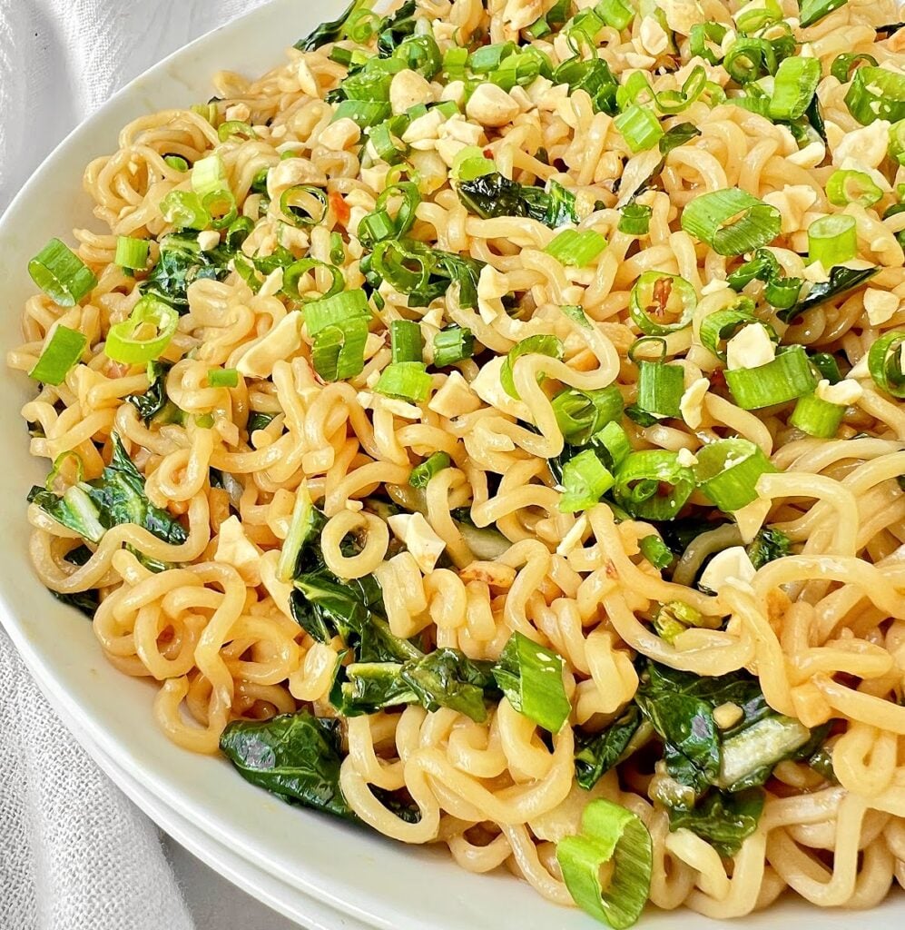 A close-up of instant ramen stir fry tossed with chopped green onions, leafy greens, and crushed peanuts. The noodles are lightly coated in sauce and garnished with fresh toppings for a flavorful meal.