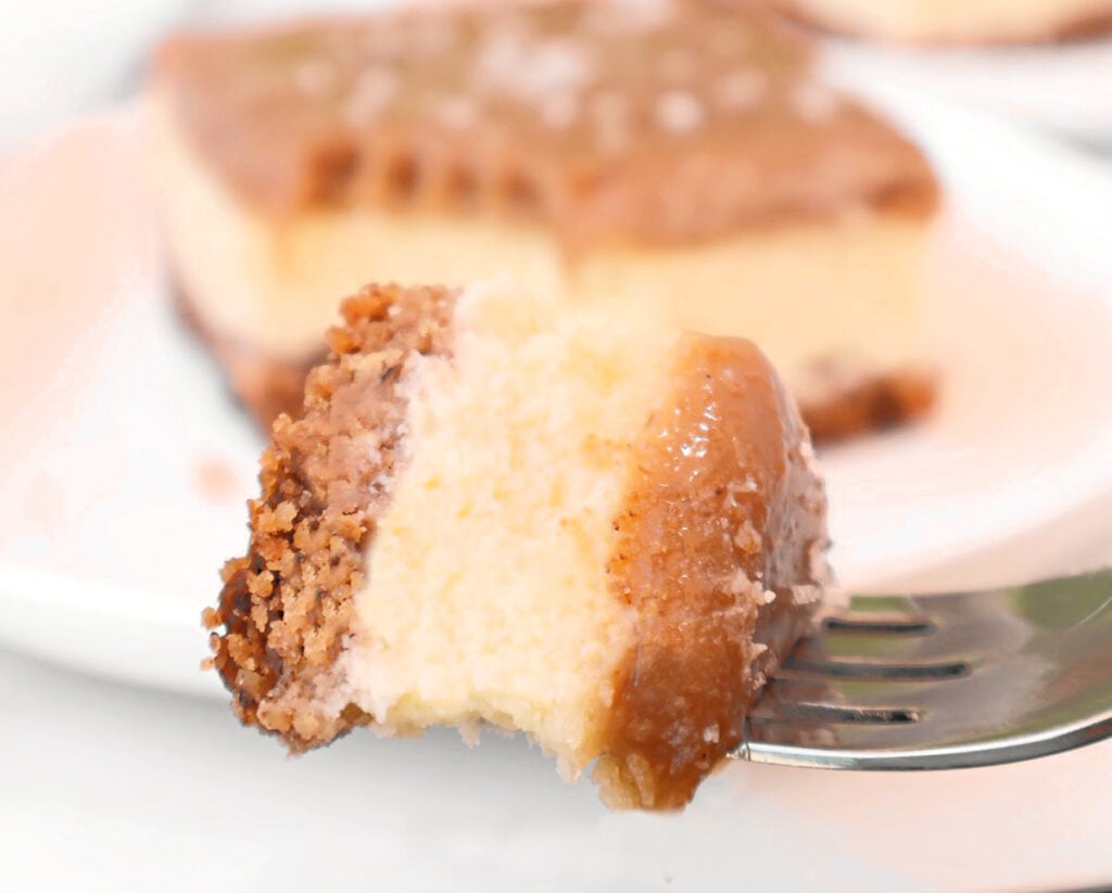 A close-up of a fork holding a bite of creamy Salted Caramel Cheesecake Bars with a crumbly crust and luscious caramel topping. A larger piece of homemade cheesecake bars is blurred in the background.