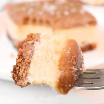 A close-up of a fork holding a bite of creamy Salted Caramel Cheesecake Bars with a crumbly crust and luscious caramel topping. A larger piece of homemade cheesecake bars is blurred in the background.