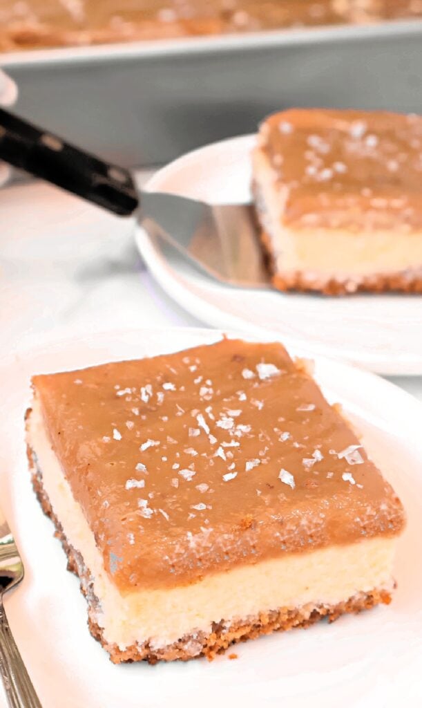 A square Salted Caramel Cheesecake Bar with a creamy middle layer, a brown caramel-like top sprinkled with flaky salt, and a crunchy base sits on a white plate. Another piece of these homemade cheesecake bars and a pan are in the background.