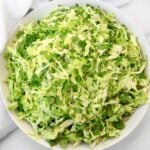A large white bowl filled with finely shredded green cabbage and cilantro, placed on a white surface with part of a white cloth visible in the background.